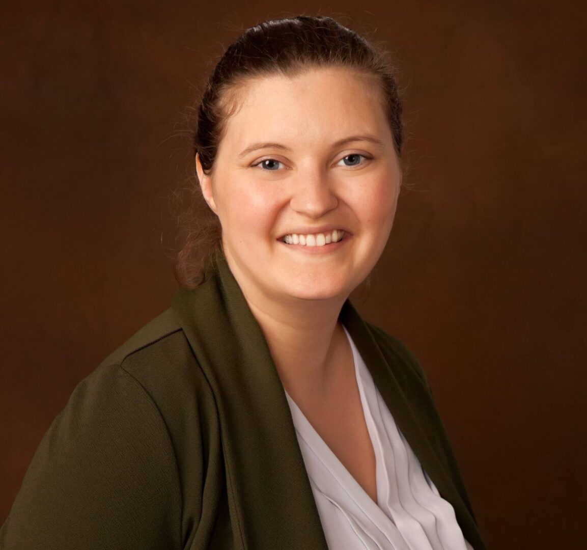 A smiling woman in a white blouse and olive green jacket against a brown background.