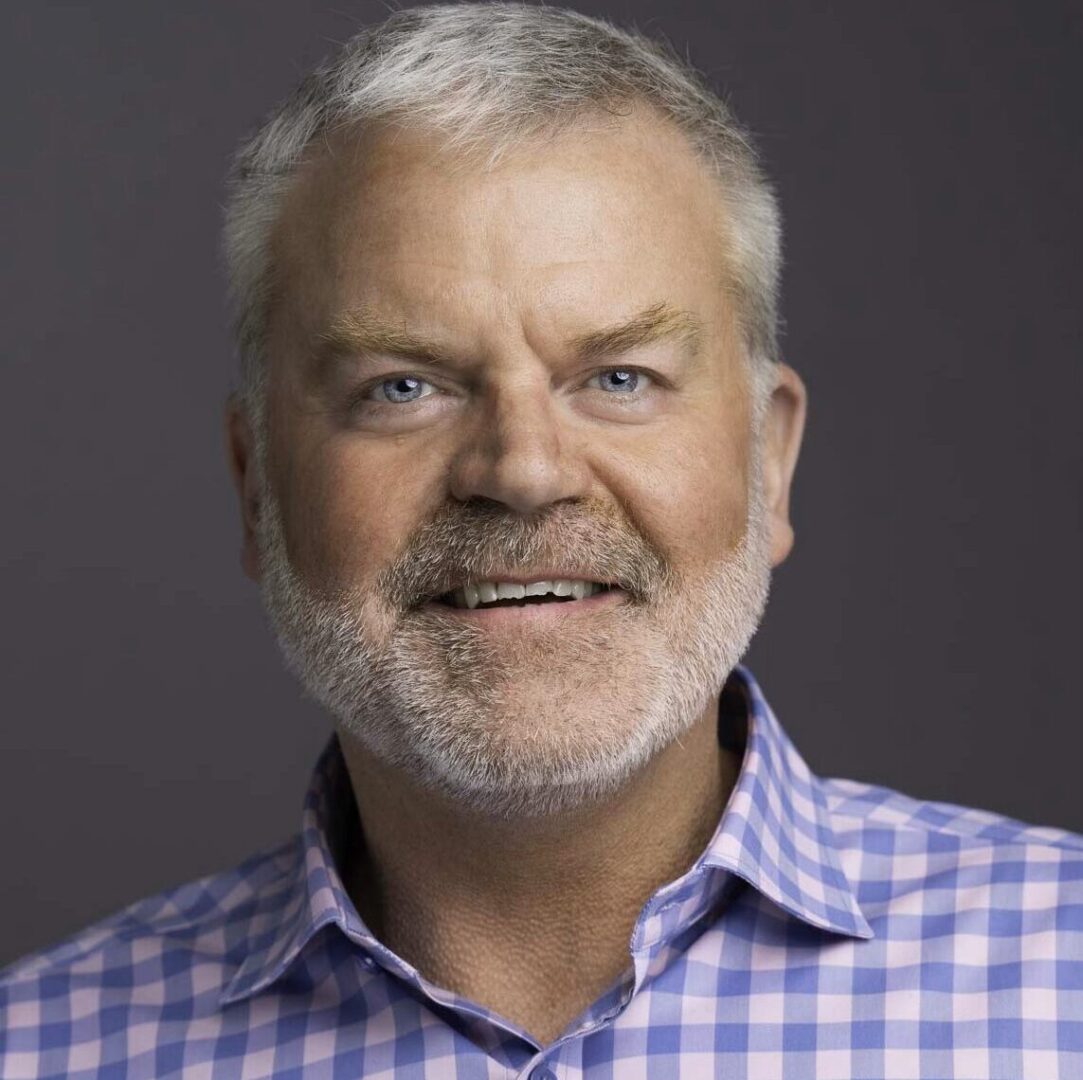 Portrait of a smiling middle-aged man with gray hair and beard in a checkered shirt.